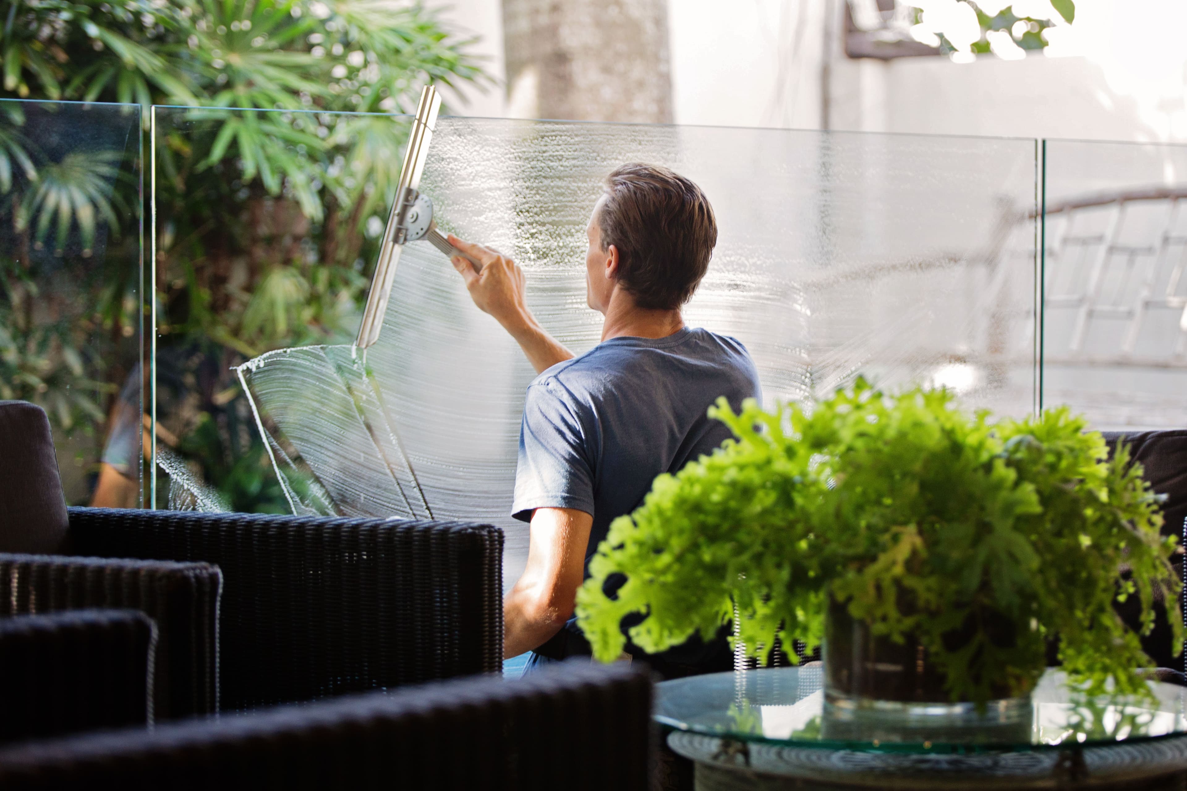 Man cleaning a glass railing with a squeegee on a patio surrounded by lush greenery.